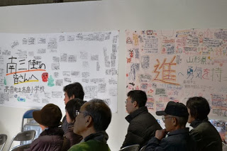 壁に「南三陸の皆さんへ」や「進～勇気を持て」などの応援メッセージが貼られた会場で、椅子に座って話を聞いている人々を写した写真