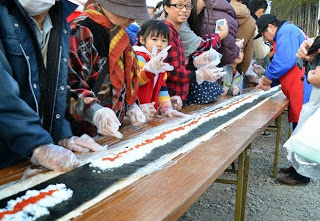 テーブルいっぱいに広げられた海苔の上にご飯といくらをのせて、参加者たちが協力して長い巻き寿司を作っている様子を写した写真