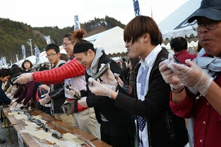 ビニール手袋をつけた多くの参加者が、完成した長い海苔巻きを一口サイズにカットしたものを手に取り、食べ始めている様子を写した写真