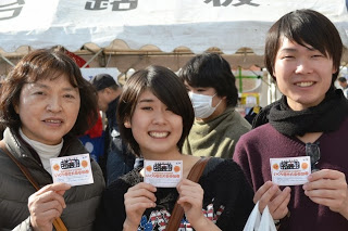 屋台の前で、イベントの抽選券を手に持って笑顔で記念撮影する三人の来場者を写した写真
