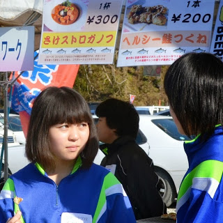 青と緑のジャージ着た女子学生二人が、「さけストロガノフ」や「ヘルシー鮭つくね」などのメニューが掲げられた屋台の前で手伝いをしている写真