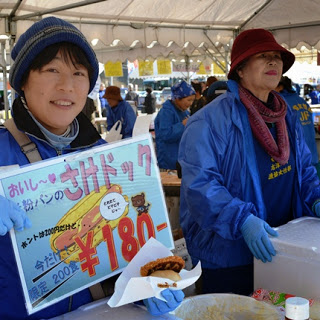 青いジャンパーを着た女性が、手作りの「さけドッグ」の看板と商品を手に笑顔で紹介している写真