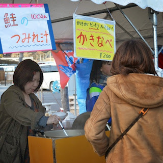 「鮭のつみれ汁」や「かきなべ」のメニューの書かれた紙が掲示されている屋台で女性が汁物を鍋からをよそって提供している様子を写した写真