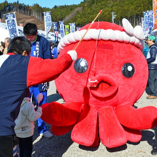 大きな赤いタコのオクトパス君の着ぐるみが会場内を歩き回り、子どもが釣り竿のような道具でオクトパス君に何かを引っかけて遊んでいる様子を写した写真