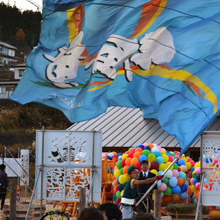 大きな青い応援旗が風に大きくなびく中、たくさんのカラフルな風船が置かれている祭り会場を写した写真