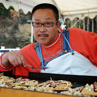 赤いシャツに白いエプロンを着た男性が鉄板の前で焼き物を仕上げながら、カメラに笑顔を向けてポーズしている様子を写した写真
