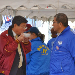 屋台の下で佐藤町長と青いジャンパーを着た関係者の男性が話をしており、佐藤町長が温かい料理を食べている様子を写した写真