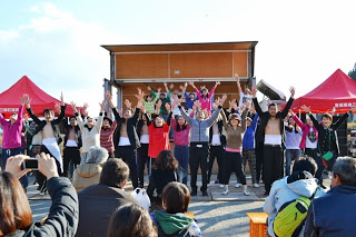 仮設ステージ前で、多くの若者や子どもたちが手を高く上げながら元気よくダンスを披露し、観客が座って見守っている様子を写した写真