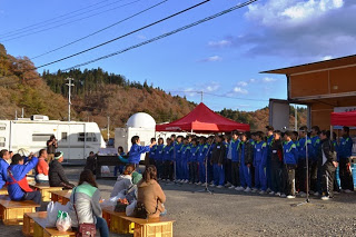 青いジャージを着た中高生の合唱団が屋外ステージ前に整列して合唱を披露しており、観客がパイプ椅子や木箱に座ってその歌声に耳を傾けている様子を写した写真