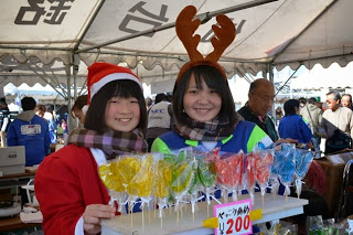 サンタ帽とトナカイのカチューシャをつけた女子学生が、カラフルなペロペロキャンディを並べて笑顔で販売している写真