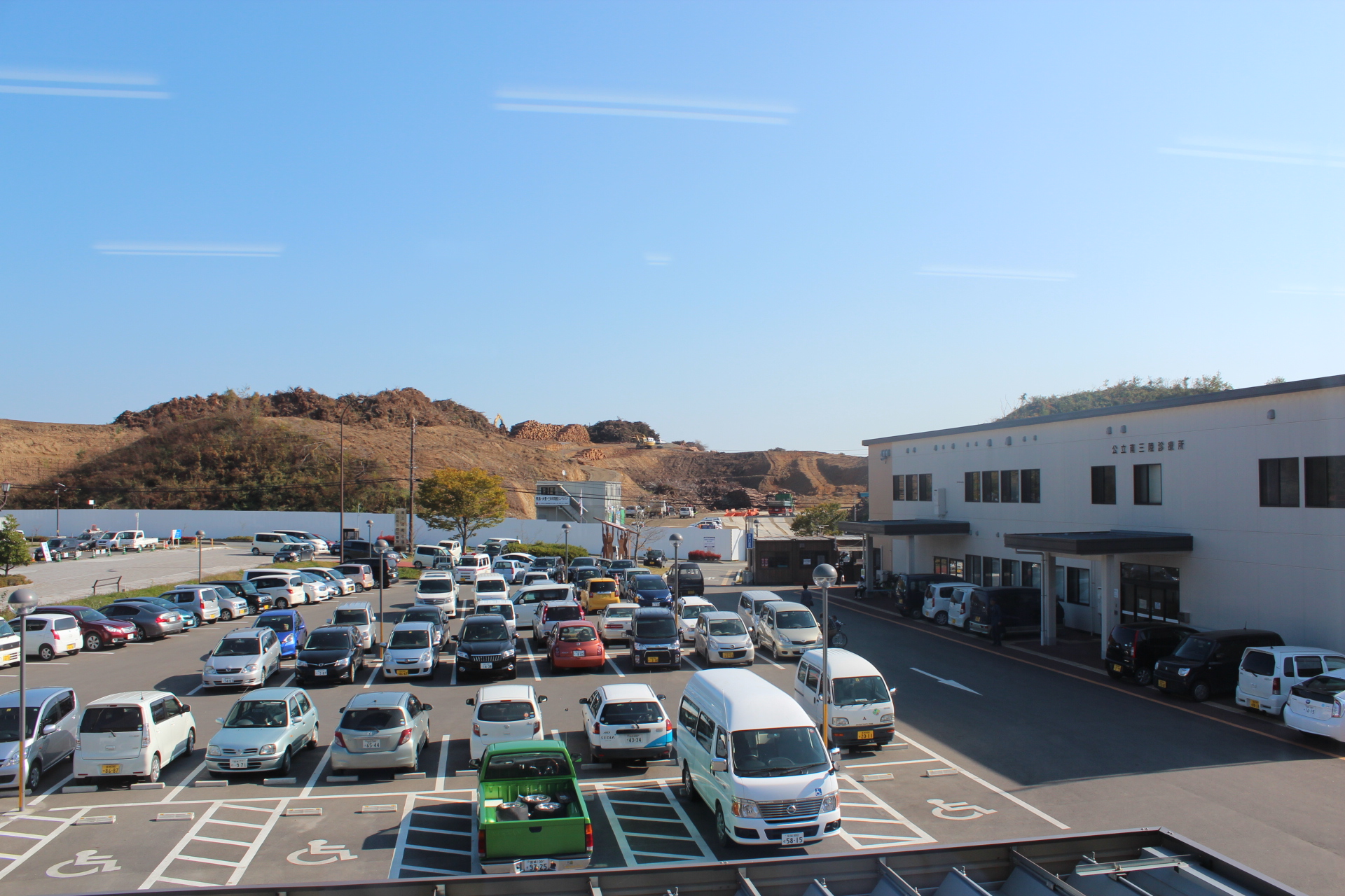 診療所の建物と沢山の車が駐車している駐車場の奥に木が伐採され積み上げられている造成地が広がっている写真