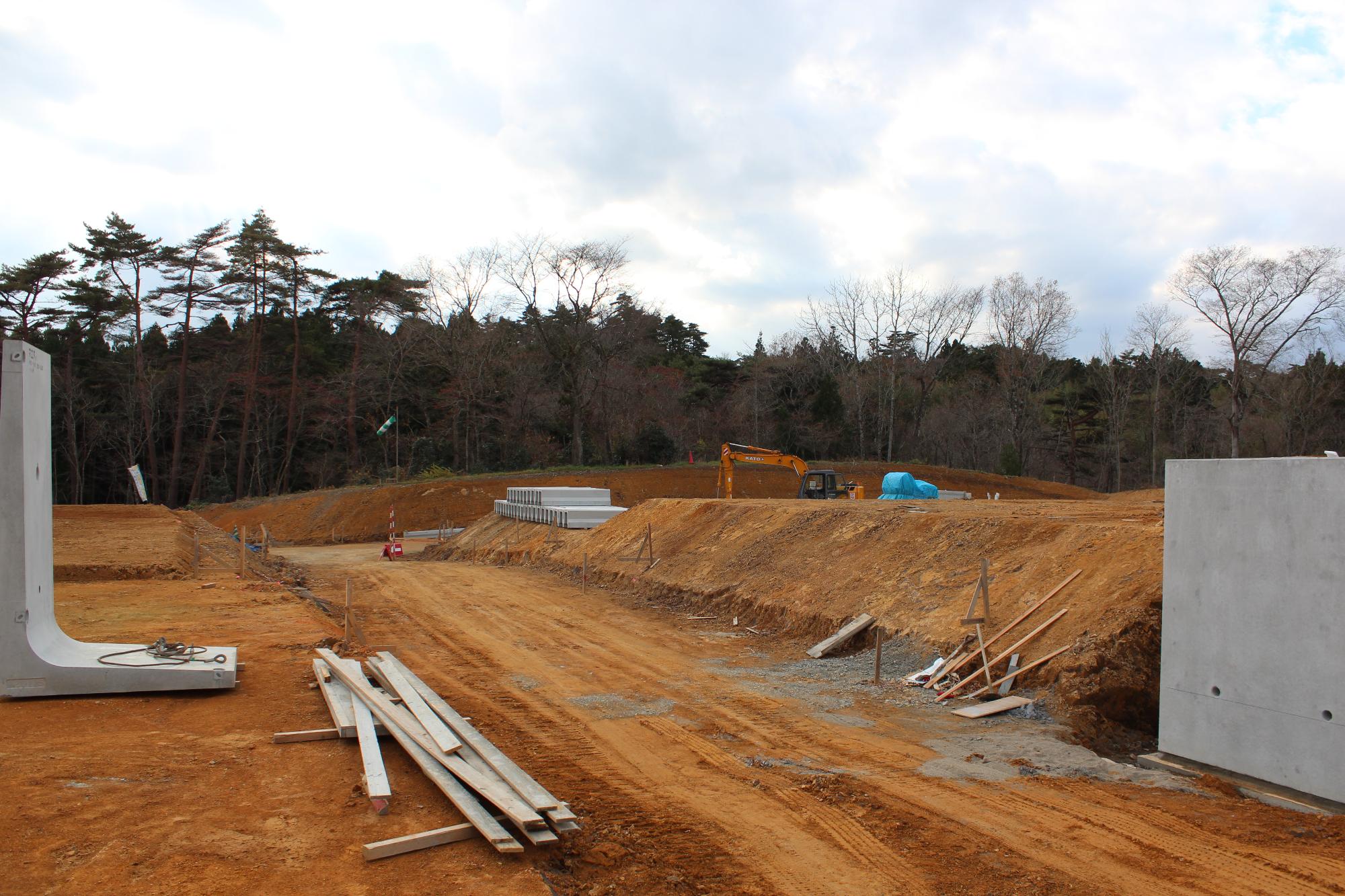 重機やコンクリート部材が点在する広い造成現場で、木々に囲まれた中に重機が作業を行っている土地開発中の写真