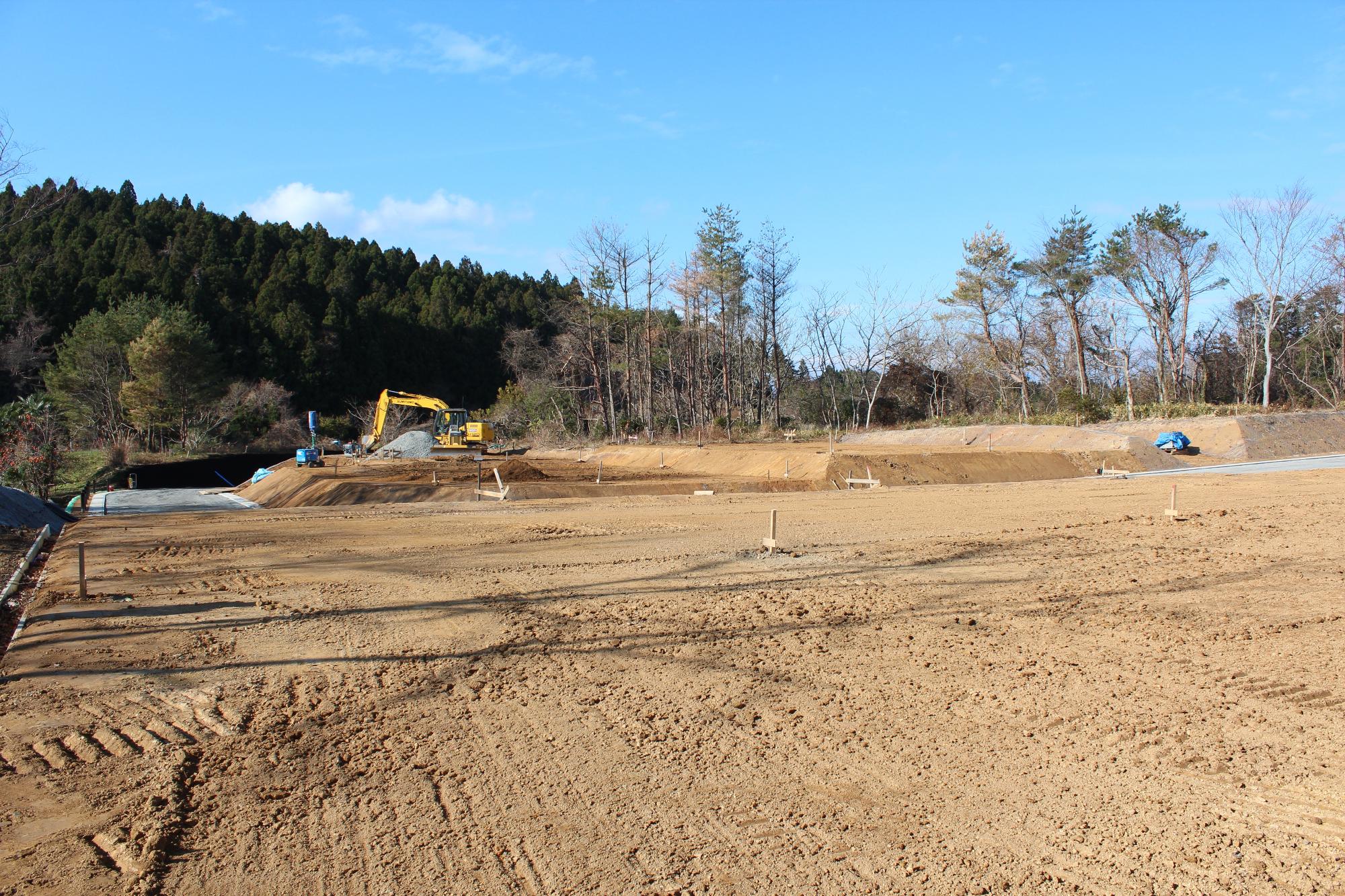 森に囲まれた広い造成地で、手前には整地された土の地面が広がり、奥では黄色いショベルカーが砂利を積み上げる作業をしている様子を写した写真