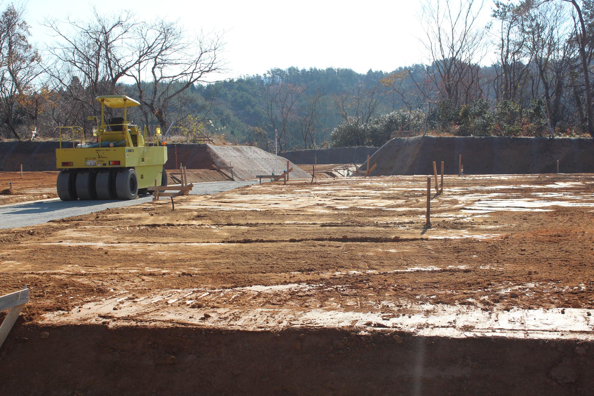 木々に囲まれた山間の開発現場で、整地された赤土の広がる地面に測量用の木杭が並び、奥には黄色いロードローラーが舗装中の道に停まっている写真