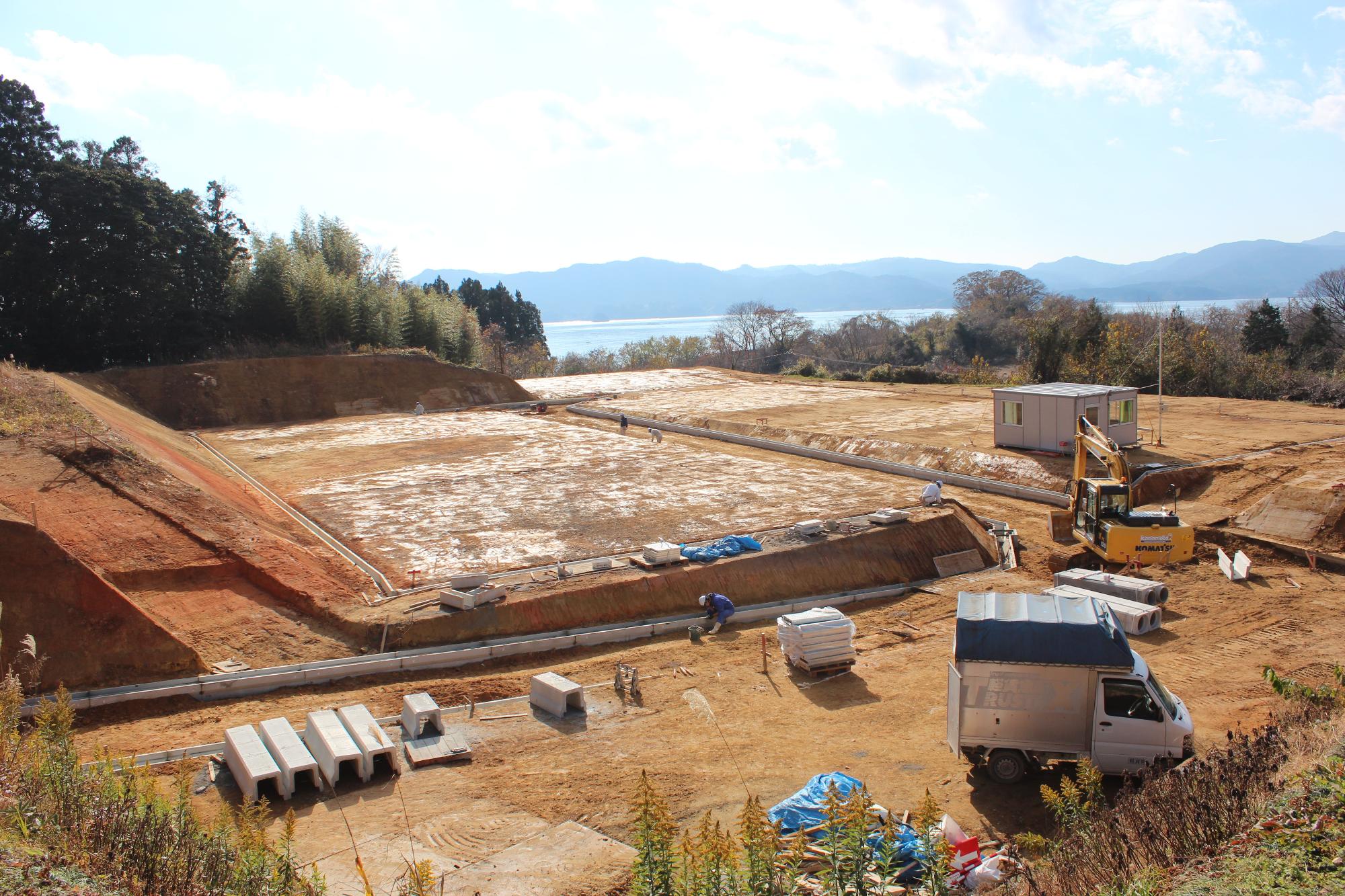 造成工事中の広い敷地に重機やトラック、作業員が点在し、遠くに海と山が見渡せる晴天の建設現場の写真