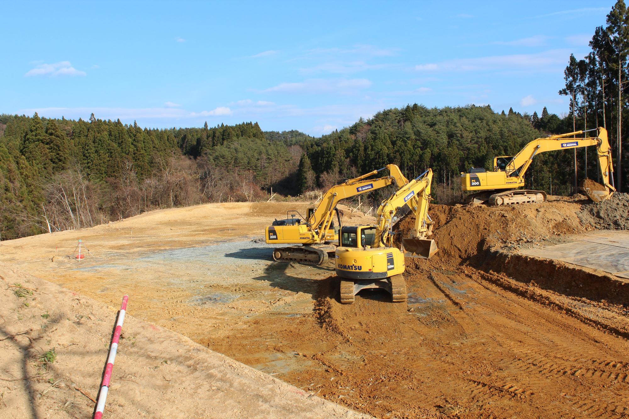 森林に囲まれた土地で、3台の黄色いショベルカーが整地作業をしている工事現場の写真