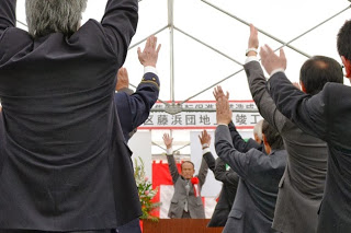 屋外のテント内で、中央の壇上に立つ男性が両手を挙げ、会場内のスーツを着た参加者たちも両手を挙げて万歳三唱をしている竣工式の写真