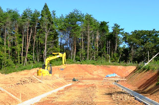 林の手前に黄色いショベルカーが停まっていて、周囲には土が掘り返された跡や資材が見られ、木々に囲まれた場所で作業が行われている工事現場の写真