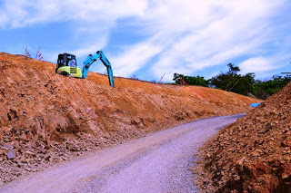 事用のブルーのショベルカーが斜面の土を掘削しており、整備中の未舗装の山道が写っている土木工事の現場の写真