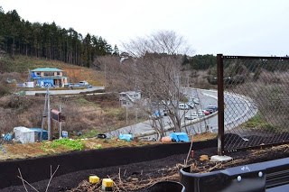 カーブした車道の脇に建設中の建物や作業用の車両が点在し、周囲には木々や斜面が広がる中、造成工事が進められている団地用地の様子を高台から見下ろしている写真