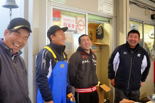 セリ場の事務所の前で、作業着や防寒着を着た漁業関係者と見られる男性4人が楽しそうに笑顔を見せている初セリの様子を写した写真
