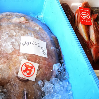 青いコンテナの中に氷と一緒に大きなアンコウが入っており、その上に「万」の赤いセリ札と手書きの値札が貼られ、隣には赤い魚が並ぶ別の発泡スチロール箱も写っているアンコウと魚の写真