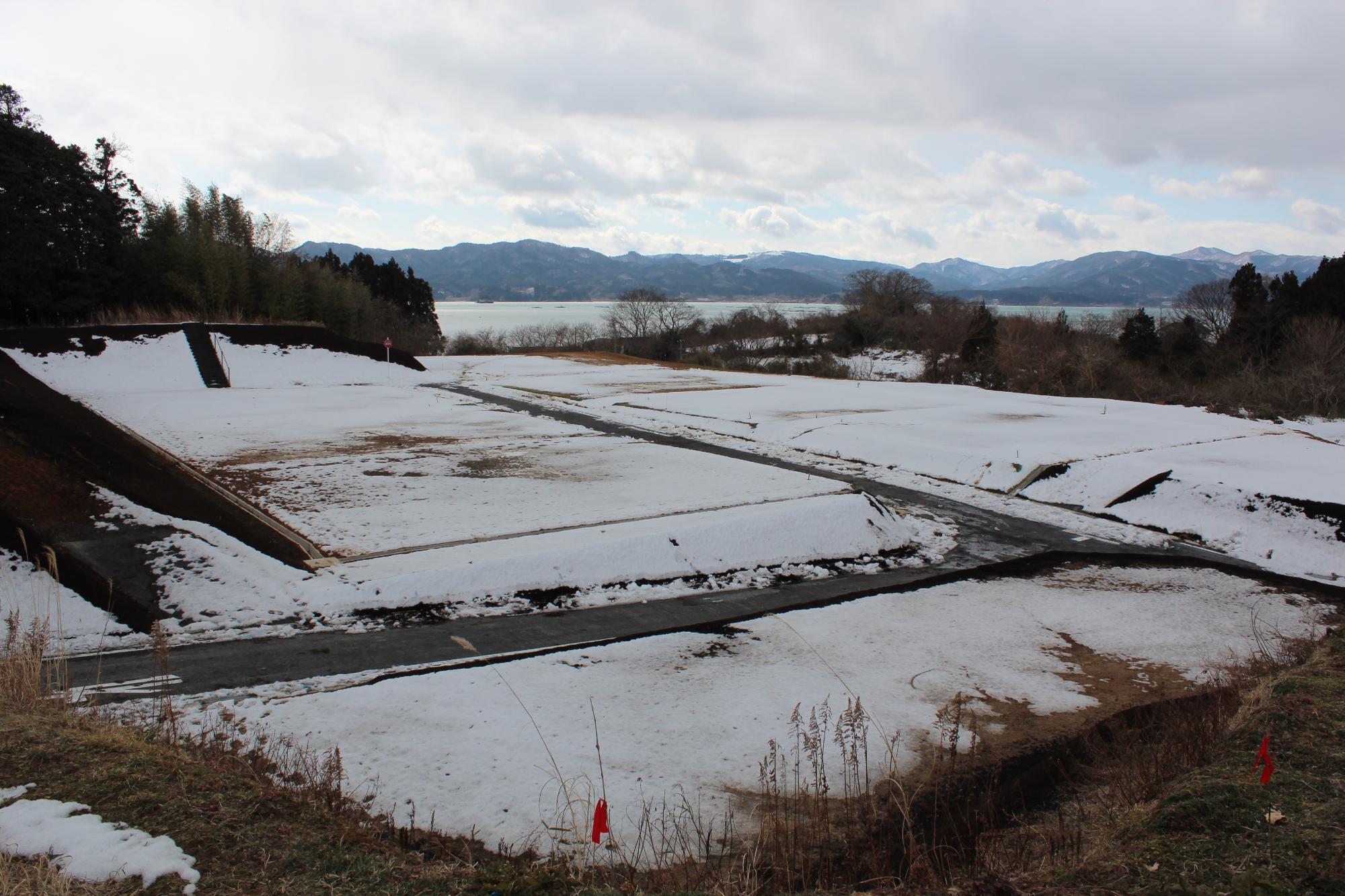 雪が残る住宅用地で、舗装された道路と造成地の境がわかるように雪の白と土の茶が対比している、海と山を背景にした写真