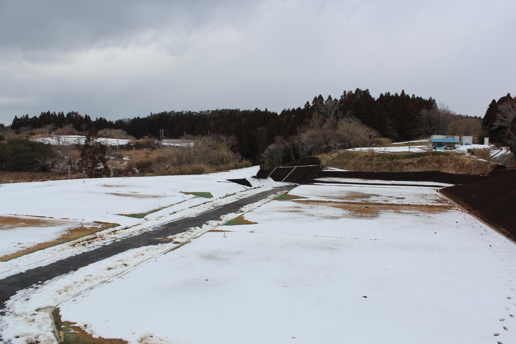 雪が積もった広い造成地で、舗装された道路が雪の中に走っており、遠くには木々と小屋のような建物が見える冬の写真