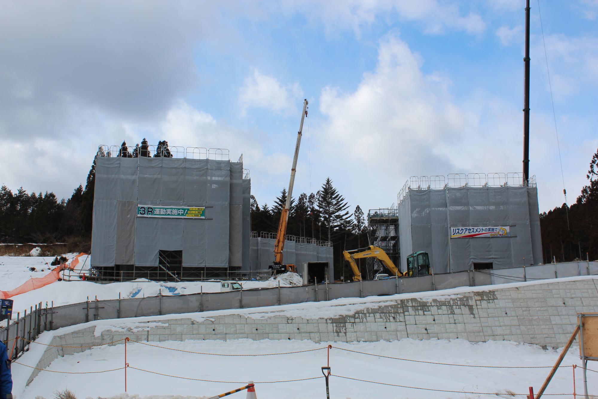 雪が積もる中、クレーン車などの重機が並ぶ建設現場で建物がシートで覆われている冬季の工事の写真