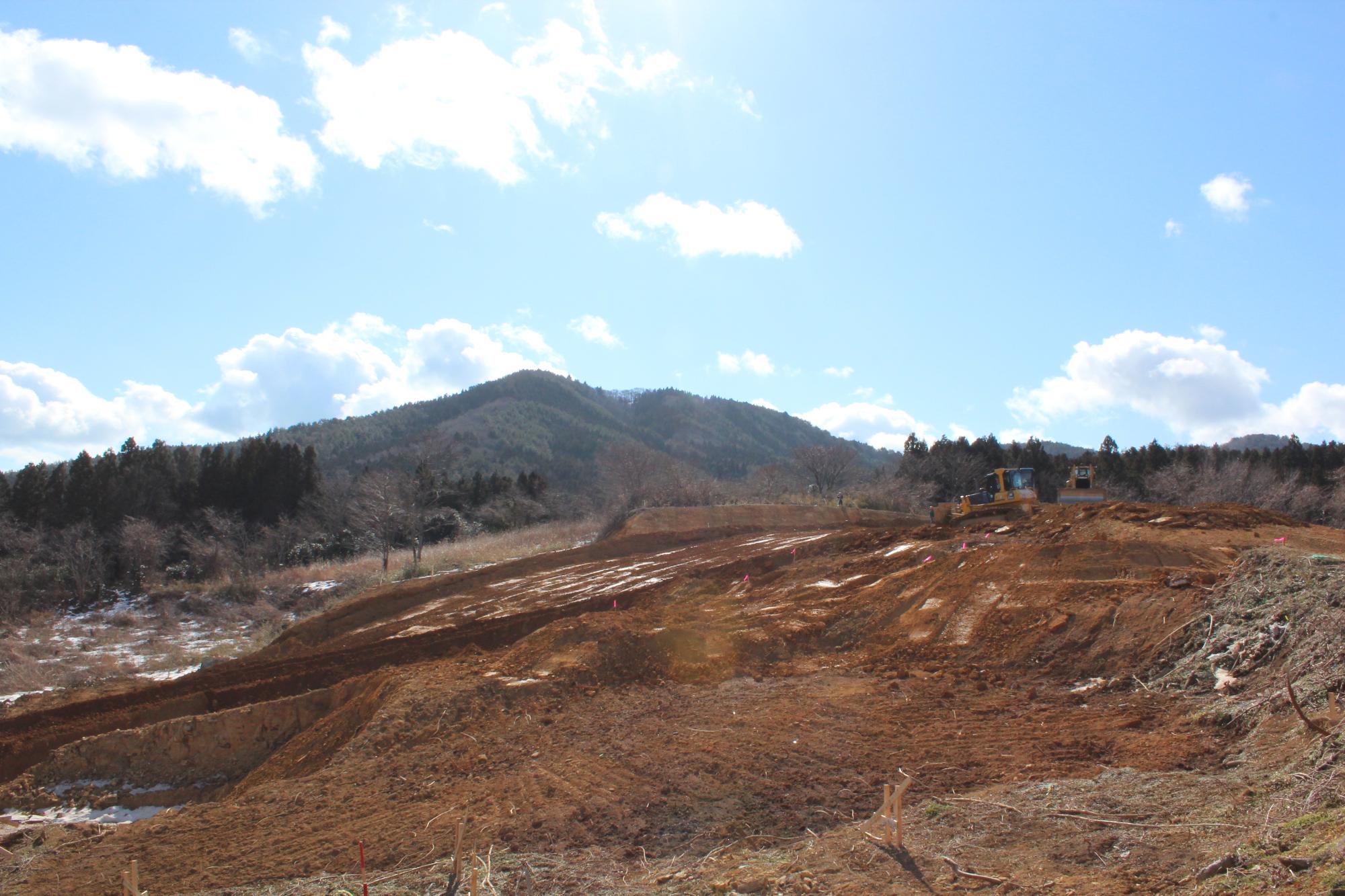 山のふもとの開けた土地で重機が整地作業をしている様子を写した写真