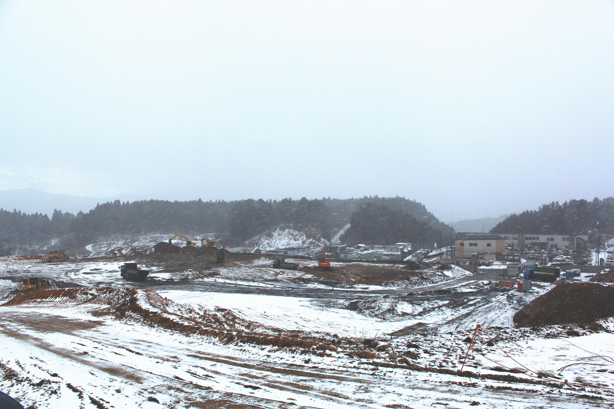 雪が積もり地面が白くなっている造成地で、ダンプカーやショベルカーなどの重機が稼働している様子の写真