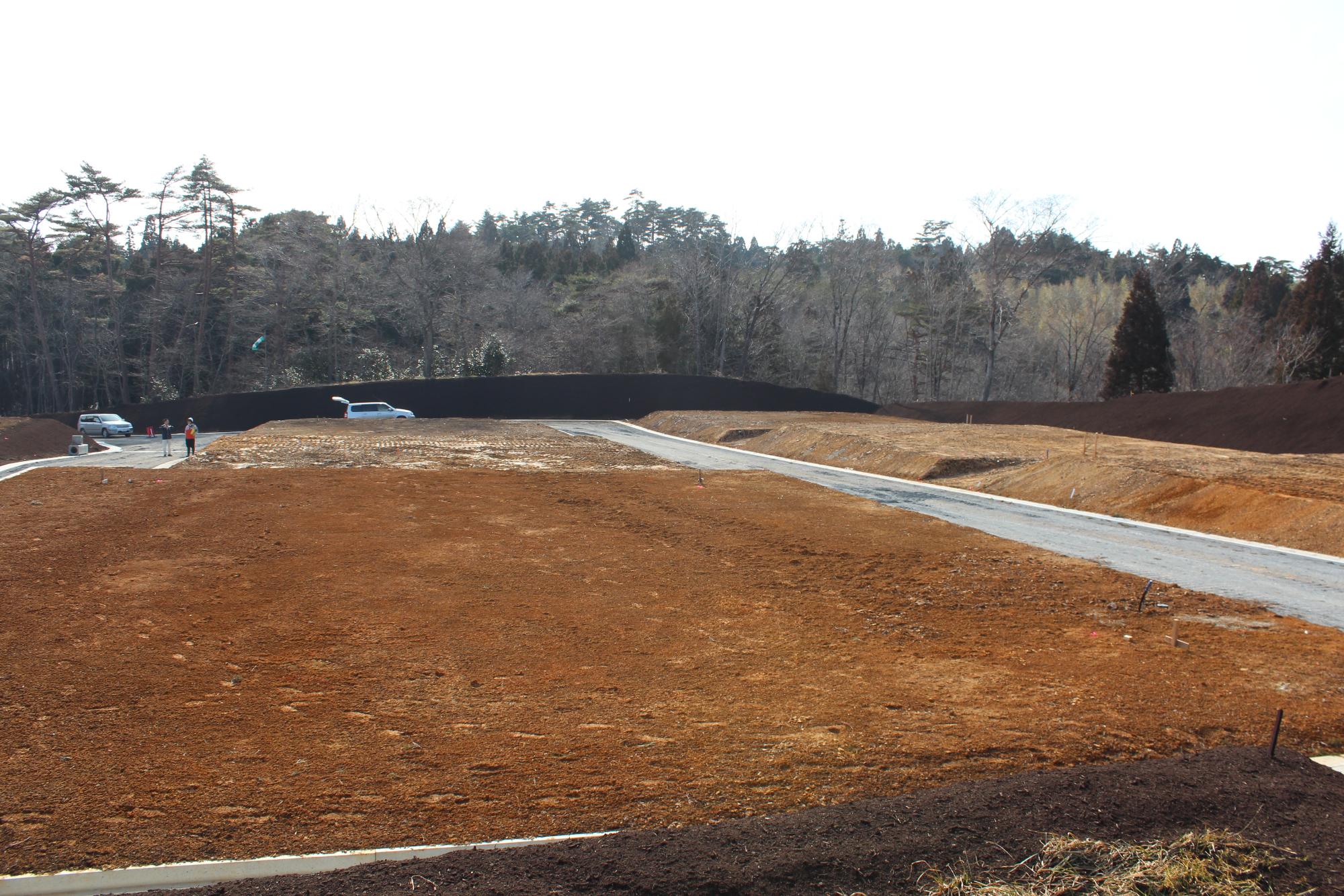 茶色い土が広がる土地に舗装されかけの道路が通り、奥に車と人が見える開発途中の住宅地の写真
