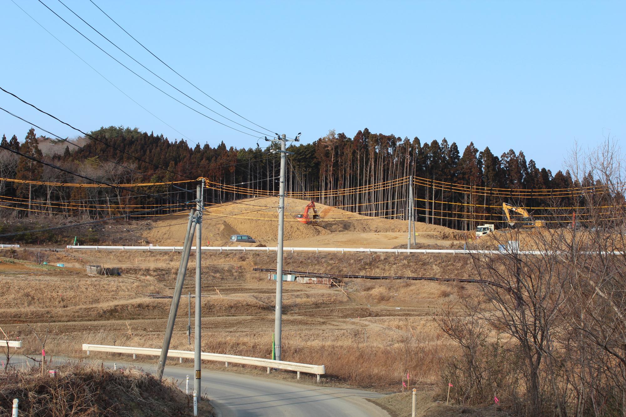 森林に囲まれた丘の造成中で、掘削機やダンプカーが土を運んでいる様子が見える開発中の斜面の写真