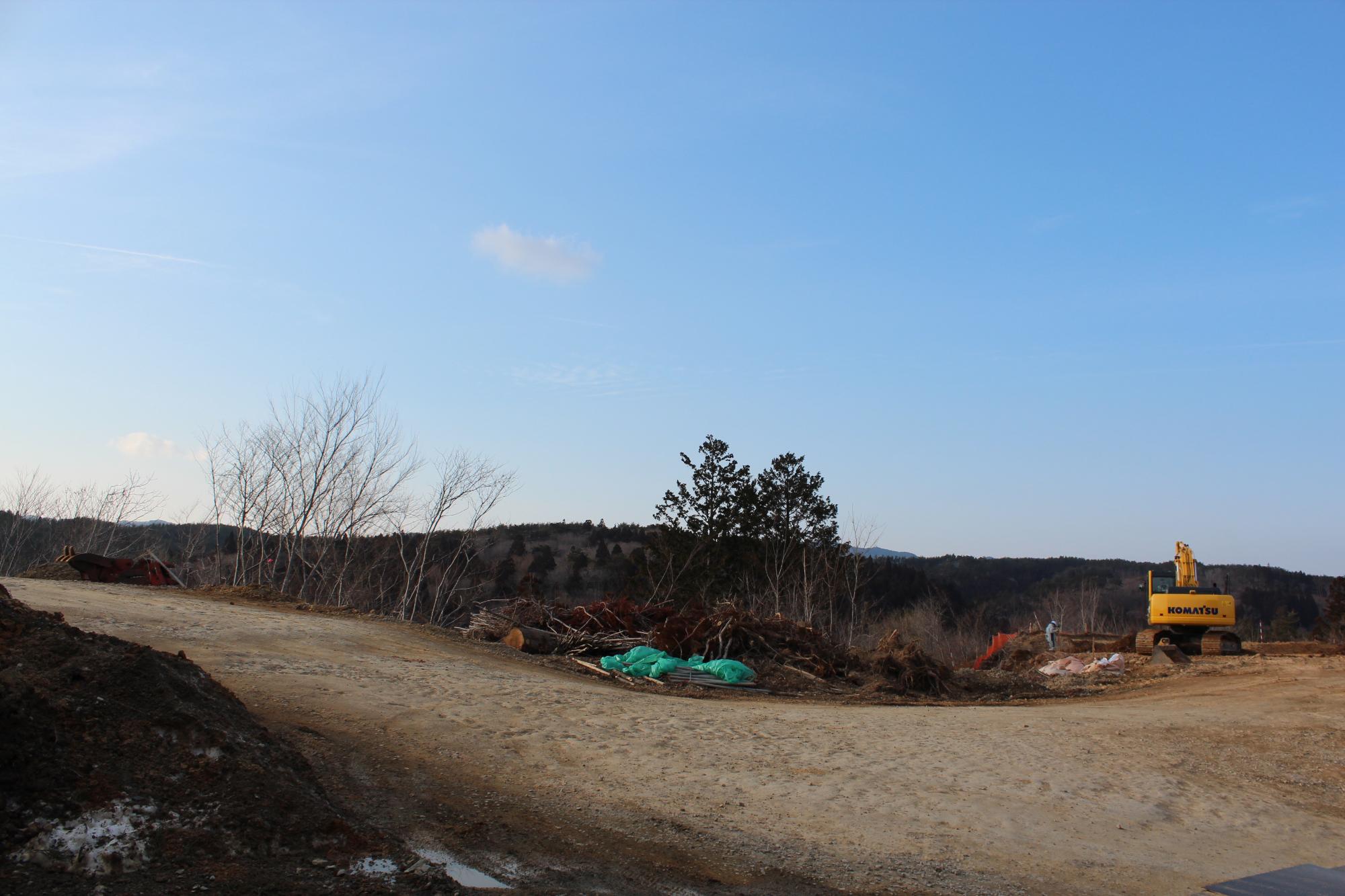 山の斜面がある造成地に伐採された木々が積まれ、重機で作業が行われている工事初期段階の写真