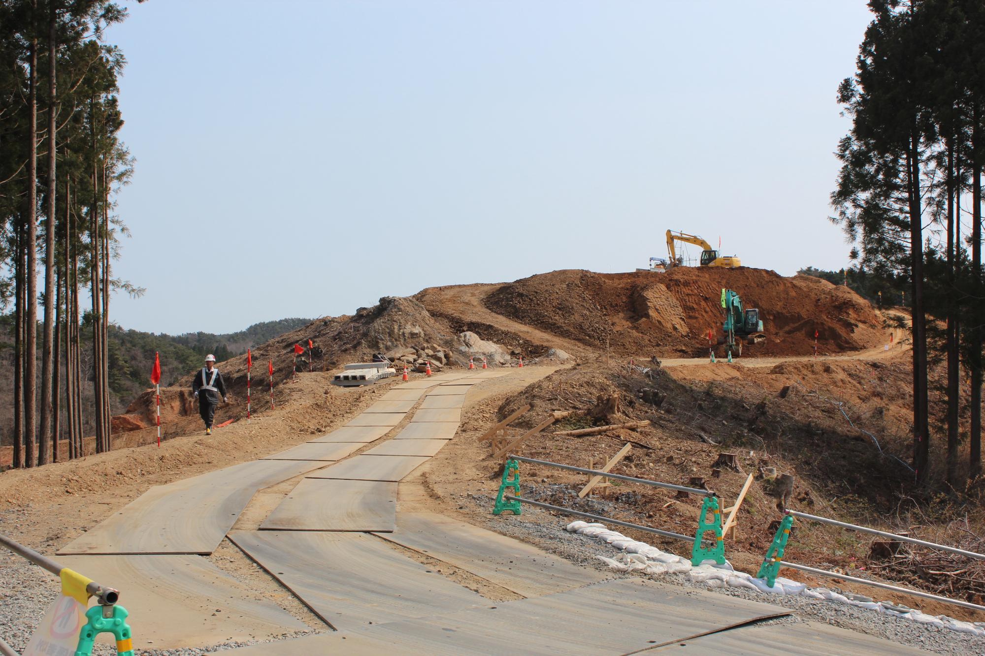 緩やかなカーブを描く仮設道路が山の斜面に沿って延び、その奥で複数の重機が造成作業を行っている工事中の写真