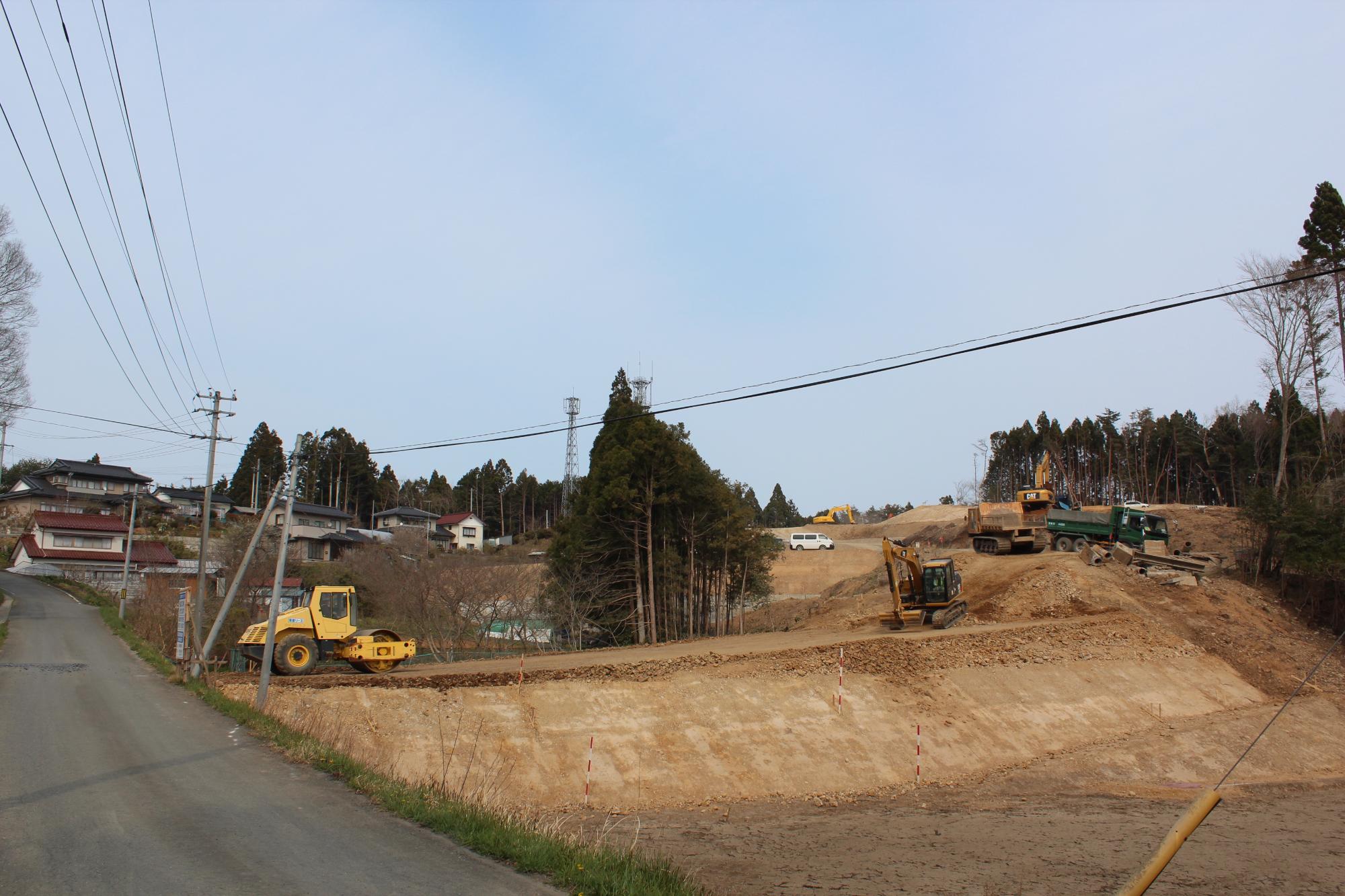 複数の重機やトラックが稼働し、傾斜地を造成している山間の工事現場の様子で、奥に住宅と通信塔が見える写真