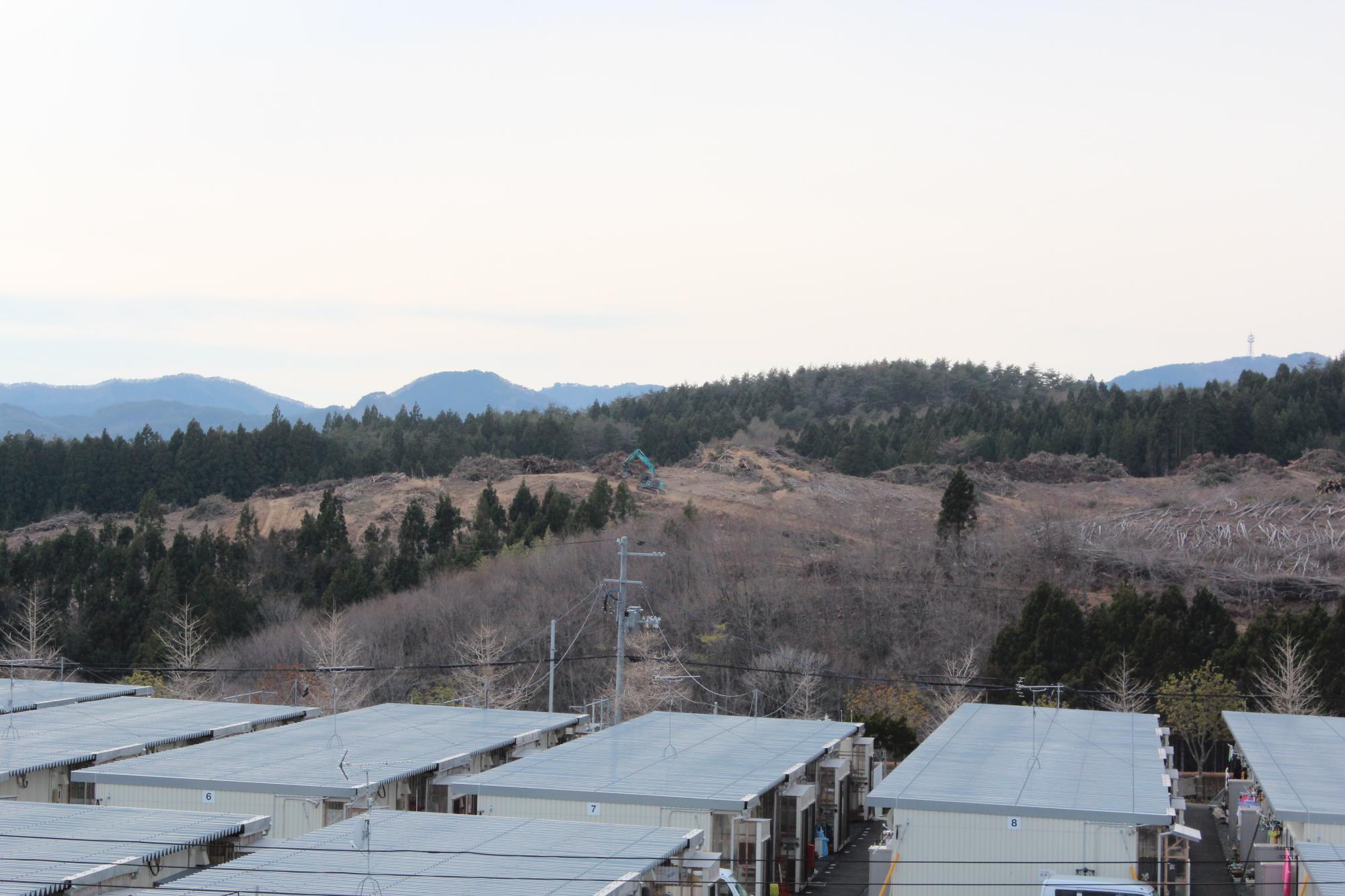 仮設住宅の上から見た木々が伐採された山の斜面と、そこに取り残された木々と重機が佇む工事現場の写真