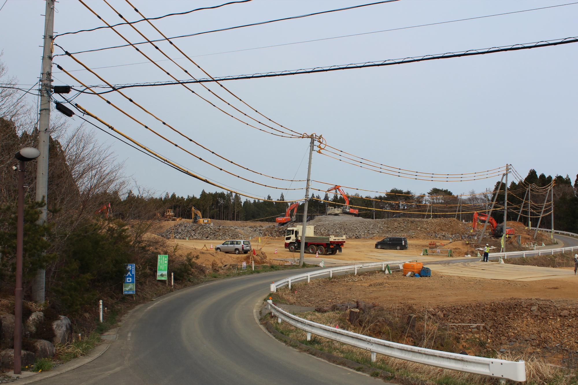 重機やトラックが岩や土を運びながら造成作業を進めている現場で、手前に曲がりくねった舗装道路と電柱に張り巡らされた電線が見え、奥には森林が広がる造成地の写真