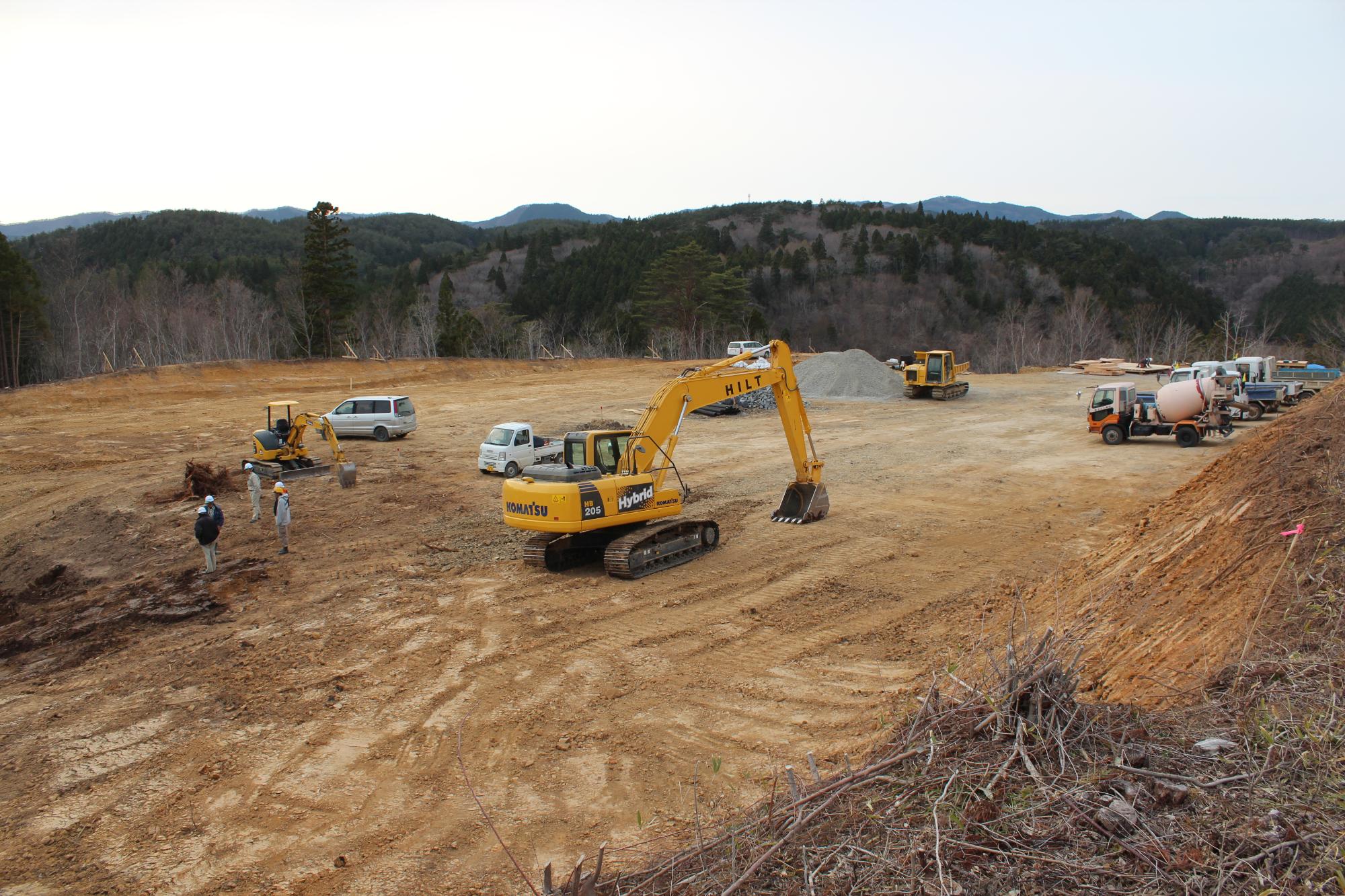 広く整地された場所で、作業員と重機、車両が配置され、山林に囲まれた中で建設準備が行われている写真