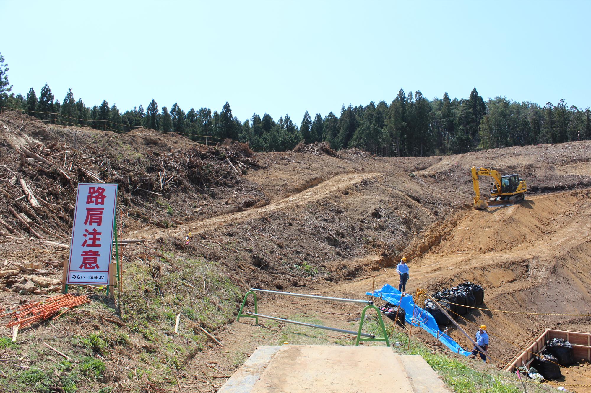 重機と作業員が整地作業を行う山の造成現場に「路肩注意」の看板が立つ写真