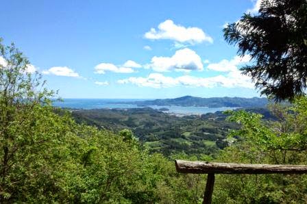木製の手すりが設置された山の上から見下ろした青い海と島々、そして広がる緑の森と空に浮かぶ雲が見える絶景の写真