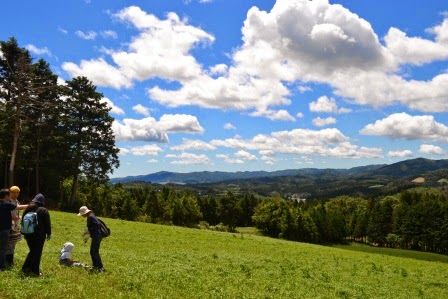 草原の中に大人と子ども数人がいて、遠くの山並みと青空、白い雲が広がっている風景の写真