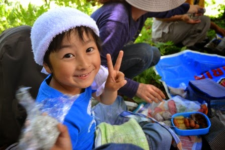 レジャーシートの上でおにぎりを手に持ちながら笑顔でピースサインをする、頭にタオルを巻いた男の子の写真