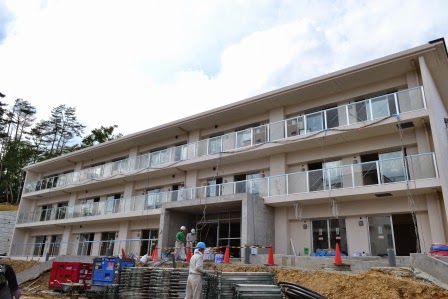 三階建ての集合住宅の正面からの様子で、ガラスの手すりがついたバルコニーが並び、建物前では作業員が作業している写真