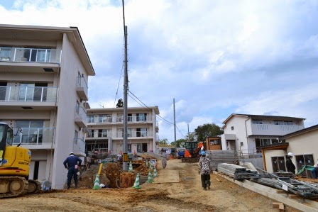 集会所と整備中の道路を隔てて、3階建ての集合住宅が傾斜地に2つ並んで建設されており、作業員が工事を進めている様子が見える写真
