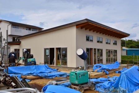 平屋建てでガラス窓が多い集会所の建物が建設中で、青いシートや資材が周囲に広がっている写真