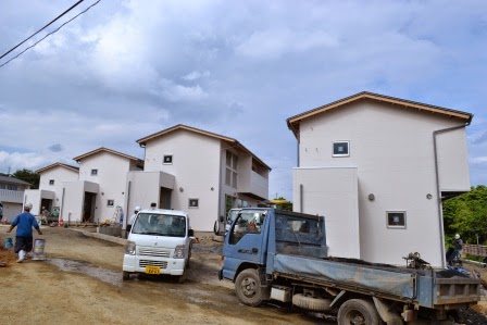 まだ整備途中の土の道路沿いに、屋根付きの白い木造戸建住宅が4棟並び、軽トラックや作業員がいる様子が見える建設現場の写真