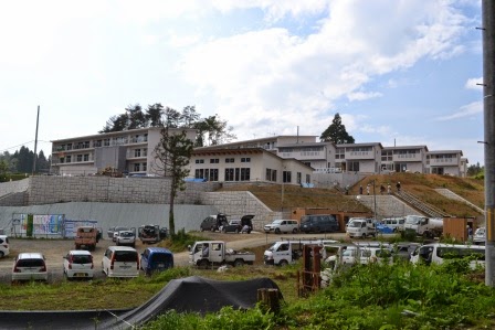 造成地の上に、緩やかな傾斜に沿って配置された集合住宅や集会所のある高台の下に多く工事の車両が停まっている写真