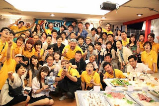 料理が並んでいるイベント会場で、参加者全員が集まってカメラに向かって笑顔でポーズをとっている集合写真