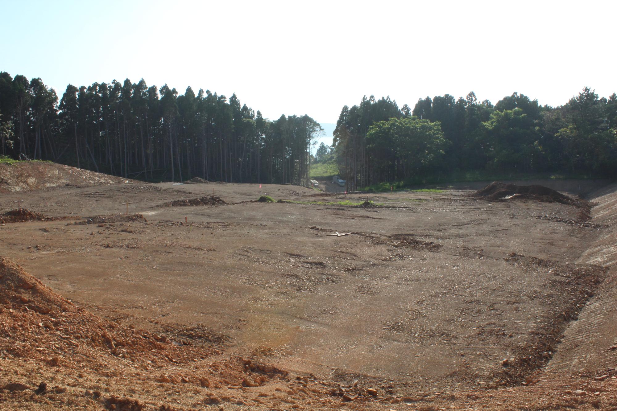 土地の整地が進み、広く開けた場所の奥に木々が生い茂る森林が見える造成工事の途中経過の写真